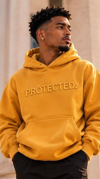 A man walking down stone steps wearing a bright yellow hoodie with 'PROTECTED.' embroidered across the chest, black cargo pants, and white sneakers with gold accents.