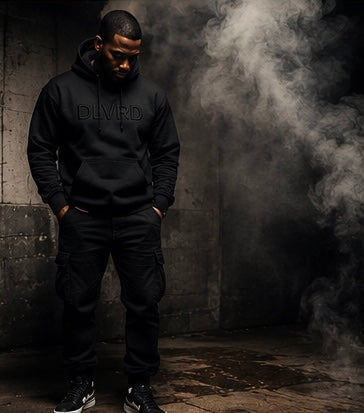 A man wearing a black pullover hoodie with 'DLVRD' printed in black across the chest, paired with black cargo pants and black sneakers, standing in a dark, smoky industrial setting.