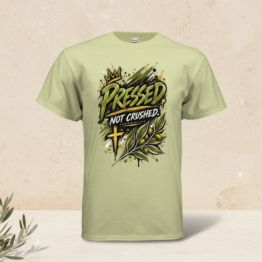 A light sage green t-shirt with a graphic print on the front that reads 'PRESSED NOT CRUSHED' in a stylized font, accompanied by a gold crown, olive branch, and cross design.