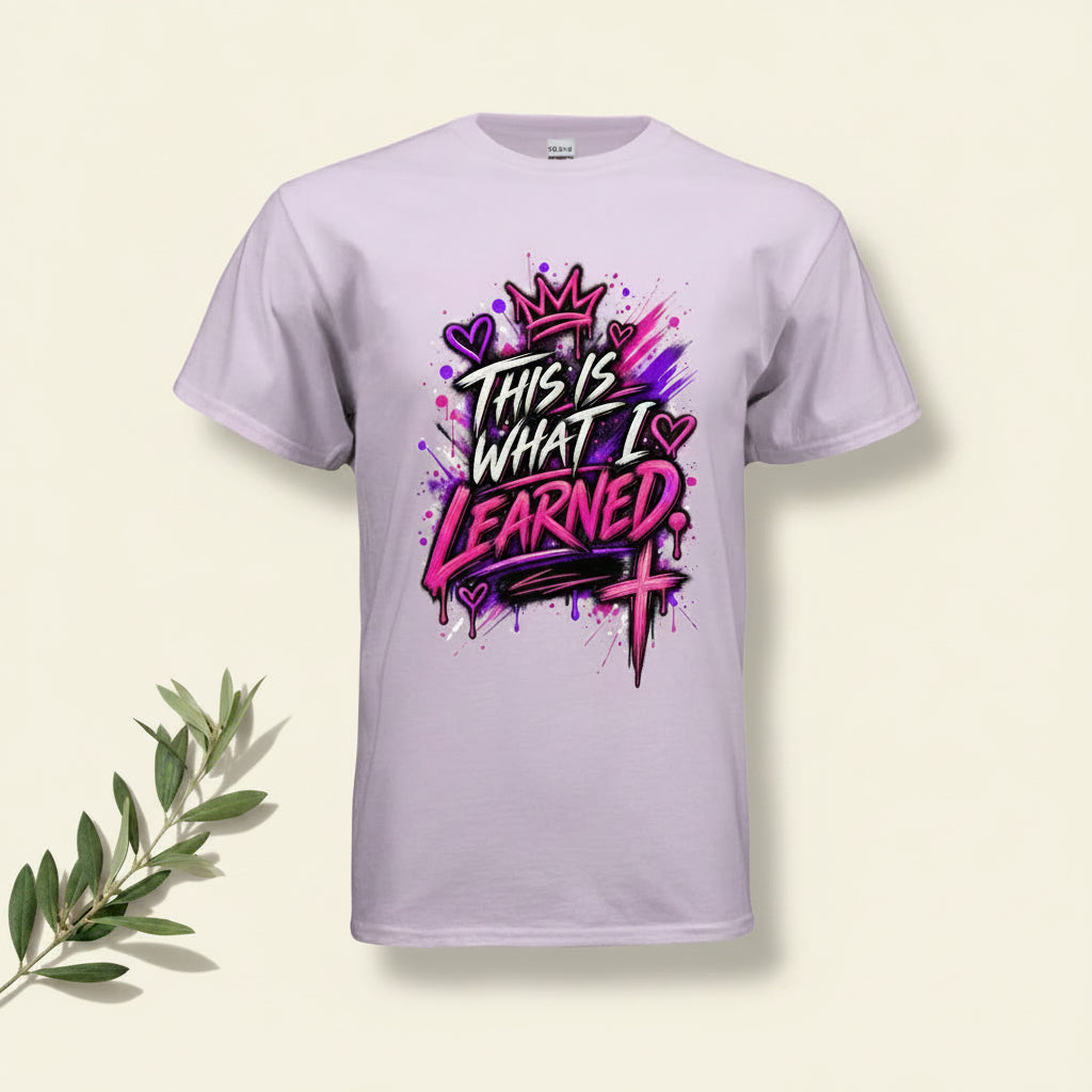 A light lavender crewneck t-shirt featuring a colorful graphic print on the front with the text 'THIS IS WHAT I LEARNED' in stylized white and pink lettering, accompanied by a crown, hearts, and a cross.