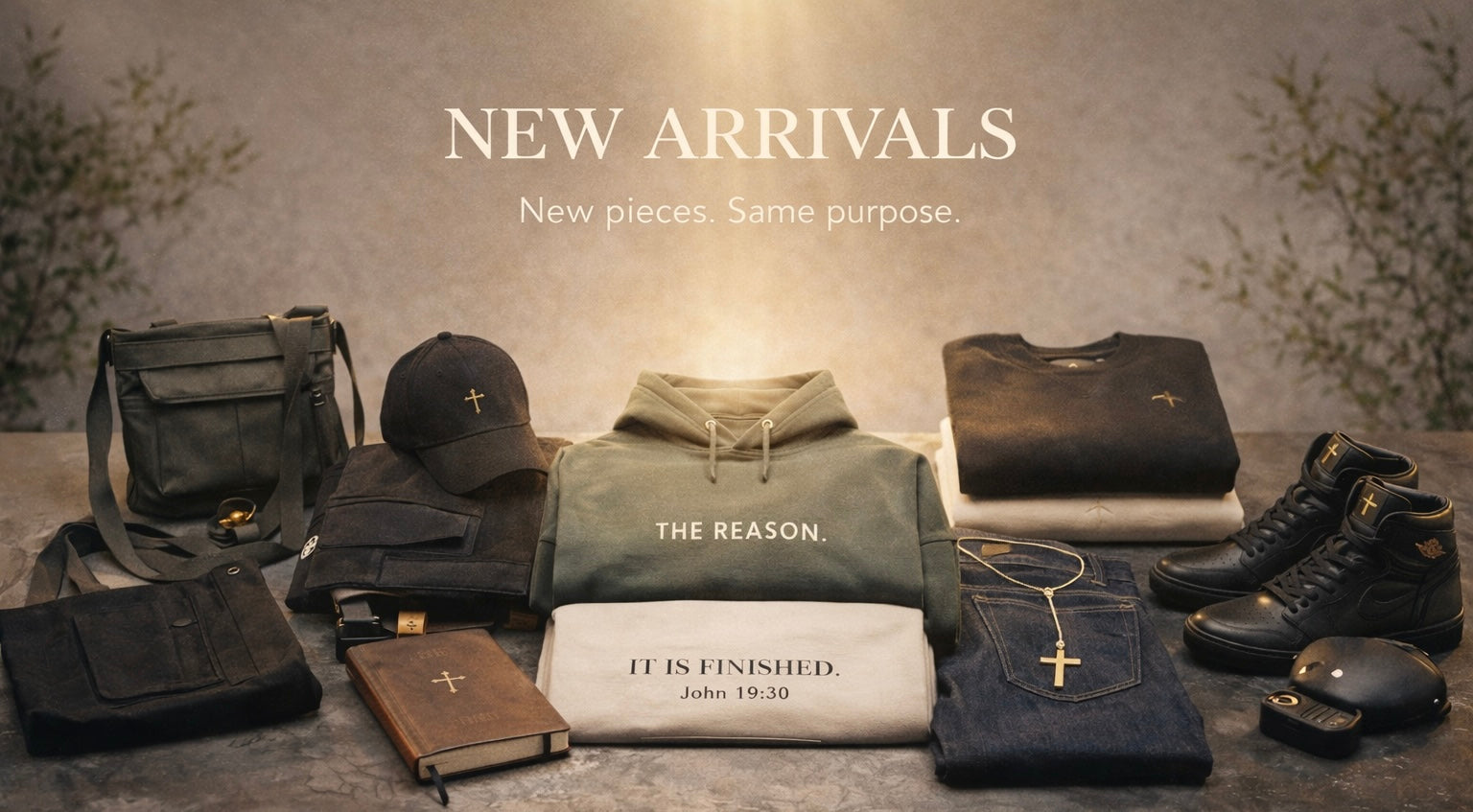 A styled product display of multiple apparel and accessory items arranged on a stone-like surface under warm lighting. Visible items include a black shoulder bag, black cap, olive green hoodie, black folded shirt, black boots, dark blue jeans, a white towel with printed text, a brown leather book or journal, and a small black pouch.