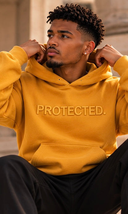 A man wearing a mustard yellow hoodie with 'PROTECTED.' embroidered on the chest, paired with black sweatpants, in an outdoor setting.