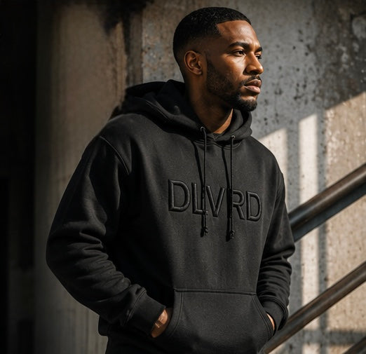 A man wearing a black pullover hoodie with 'DLVRD' printed in black across the chest, standing against a textured concrete wall with dramatic side lighting.