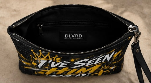 A black zippered clutch bag with a yellow and white graffiti-style graphic reading 'I'VE SEEN HIM DO IT' and a crown motif, shown open on a neutral textured beige background. The interior label reads 'DLVRD'.