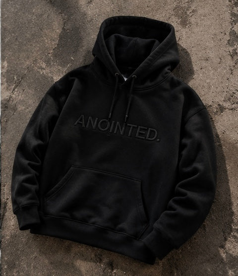 A black pullover hoodie with 'ANOINTED.' embroidered in tonal black across the chest, laid flat on a textured concrete surface.