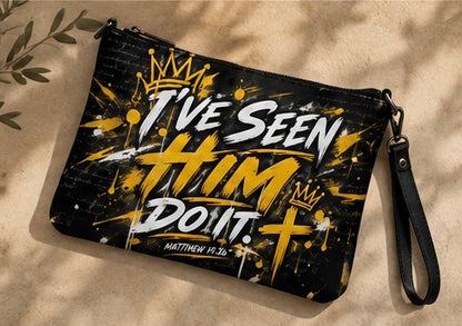 A black wristlet pouch with a yellow and white graphic print reading 'I've Seen Him Do It' and 'Matthew 18:13-14' with crowns and a cross, shown on a textured beige surface with leaf shadows.
