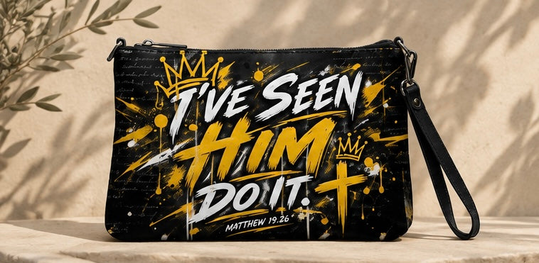 A black wristlet clutch bag with a graphic print featuring the text 'I'VE SEEN HIM DO IT. MATTHEW 19:26' in white and yellow with gold crowns and splatter details, displayed on a stone surface with leaf shadows.