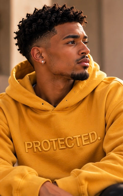 A man wearing a mustard yellow hoodie with the word 'PROTECTED.' embroidered across the chest, shown in a close-up portrait.