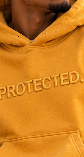 Close-up of a mustard yellow hoodie worn by a model, featuring the word 'PROTECTED.' in raised embroidery across the chest.