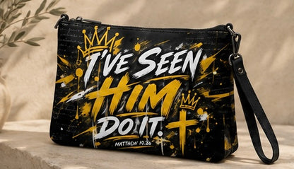 Black wristlet clutch bag with a black strap, featuring a graphic print with the text 'I'VE SEEN HIM DON'T.' in white and yellow, gold crown and cross illustrations, and 'MATTHEW 12:36' at the bottom, displayed on a stone surface.