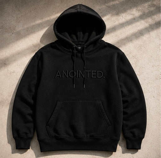 A black pullover hoodie with 'ANOINTED.' printed in tonal black across the chest, laid flat on a textured concrete surface with diagonal shadows.