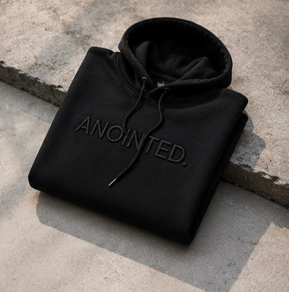 A folded black pullover hoodie with 'ANOINTED.' embroidered in black across the chest, resting on a textured concrete surface.