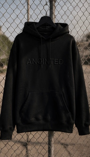 A black pullover hoodie with tonal black embroidered text reading 'ANOINTED.' across the chest, hanging on a black hanger in front of a chain-link fence.