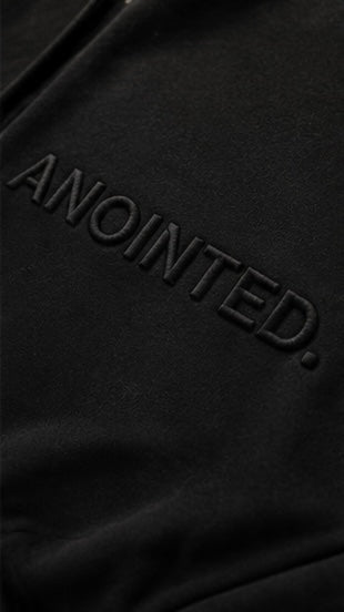 Close-up of a black fabric surface with the word 'ANOINTED.' embossed in matching black lettering.