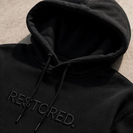 A close-up of a black pullover hoodie with 'RESTORED.' embroidered in tonal black across the chest, shown against a light textured concrete background.