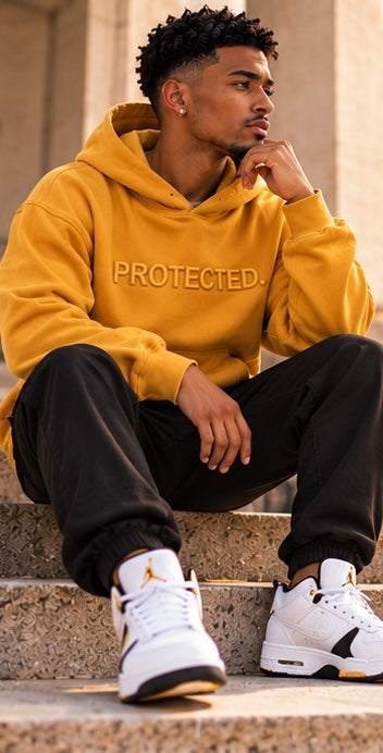 A man sitting on stone steps wearing a mustard yellow hoodie with 'PROTECTED.' printed across the chest, black sweatpants, and white sneakers with gold accents.