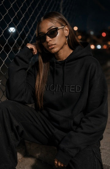 A woman wearing a black oversized hoodie with the word 'ANOINTED' in tonal black embroidery across the chest, paired with black sweatpants and dark sunglasses, sitting outdoors at night in front of a chain-link fence.
