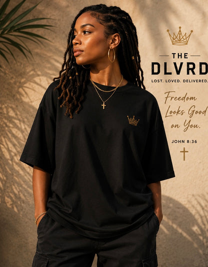 A woman wearing a black oversized T-shirt with a small gold crown graphic on the left chest, posed against a warm textured background with text on the right reading 'THE DLVRD' and 'Freedom Looks Good on You. JOHN 8:36'.