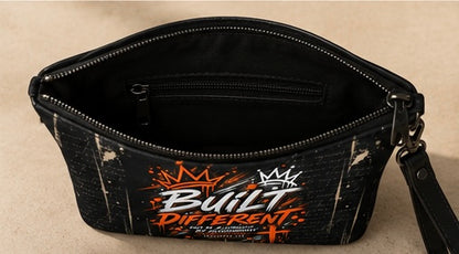 A black zippered pouch bag with a distressed graphic print featuring the words 'Built Different' in white and orange, with a crown graphic above, shown open on a beige background.
