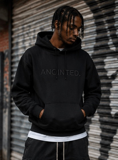 A man wearing a black pullover hoodie with 'ANOINTED.' printed in tonal black across the chest, standing in front of a closed metal shutter.