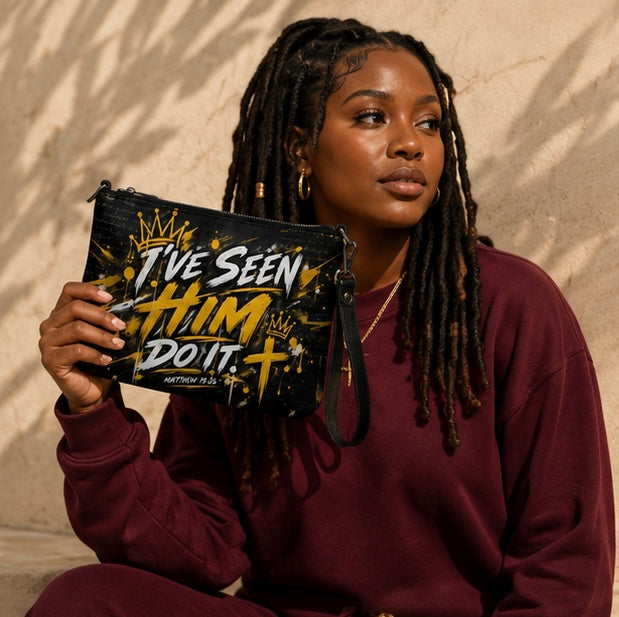 A woman with long black dreadlocks holds up a black clutch bag with a gold and white graphic that reads 'I've Seen Him Do It'. She is wearing a burgundy sweatshirt and is posed against a textured beige wall with soft shadows.