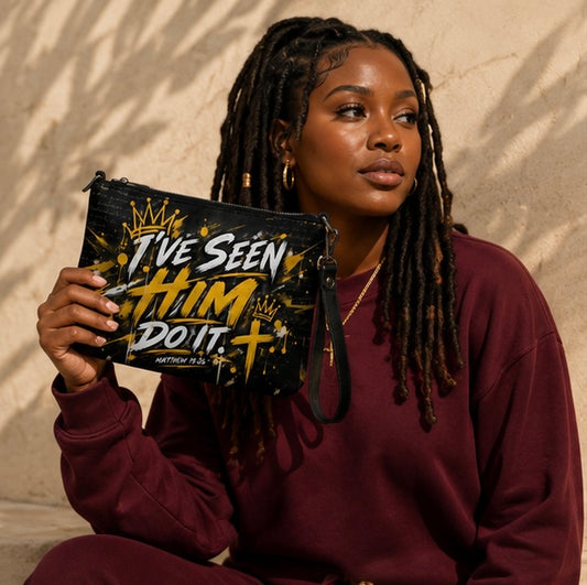 A woman with long black dreadlocks holds up a black clutch bag with a gold and white graphic that reads 'I've Seen Him Do It'. She is wearing a burgundy sweatshirt and is posed against a textured beige wall with soft shadows.