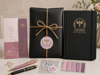 A coordinated gift set arranged on a light surface: a black wrapped package tied with twine and a round pink tag, a black notebook with gold 'The Dlyrd' logo, a pink-and-white card in a clear sleeve, two pens, a small 'Jesus is enough' card, and pink and mauve color swatches. No person or real environment is the focus, so this is Product/Packaging.