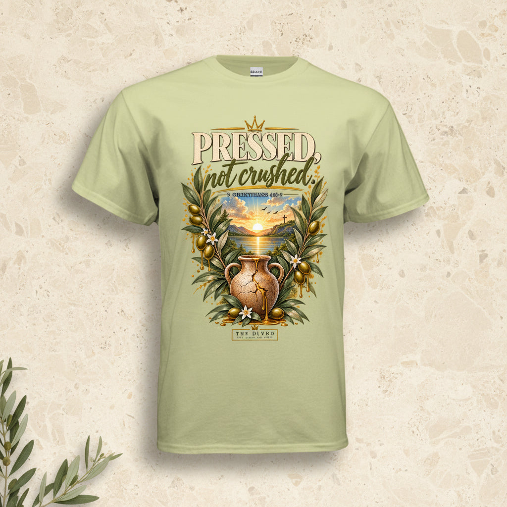 A light sage green t-shirt with a detailed graphic on the front. The graphic features the text 'PRESSED, not crushed' in a distressed serif font, above an illustration of a gold-toned amphora vase surrounded by olive branches and a sunset landscape.
