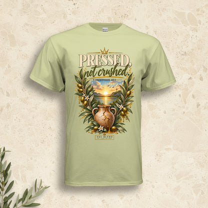 A light sage green t-shirt with a detailed graphic on the front. The graphic features the text 'PRESSED, not crushed' in a distressed serif font, above an illustration of a gold-toned amphora vase surrounded by olive branches and a sunset landscape.