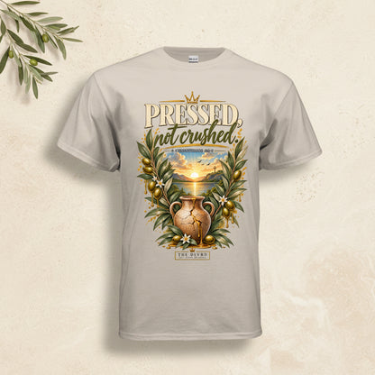 A light beige crewneck t-shirt featuring a detailed graphic print on the front. The graphic displays a cracked clay jug surrounded by olive branches with green leaves and gold olives, set against a sunset background, with the text 'PRESSED not crushed' and 'EX HIBIT'.