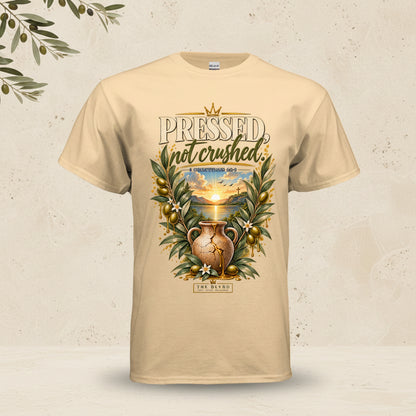 A beige crewneck t-shirt featuring a graphic print on the front with the text 'PRESSED not crushed' and an illustration of an ancient clay pot surrounded by olive branches and a sunset landscape.