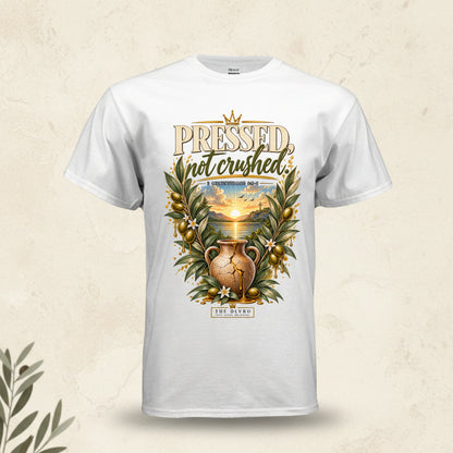 A white short-sleeve crewneck t-shirt featuring a detailed graphic print on the front. The graphic displays the words 'PRESSED not crushed' in a stylized font, surrounding an illustration of a golden sun setting over a landscape with a cracked clay pot, olive branches, and gold olives.