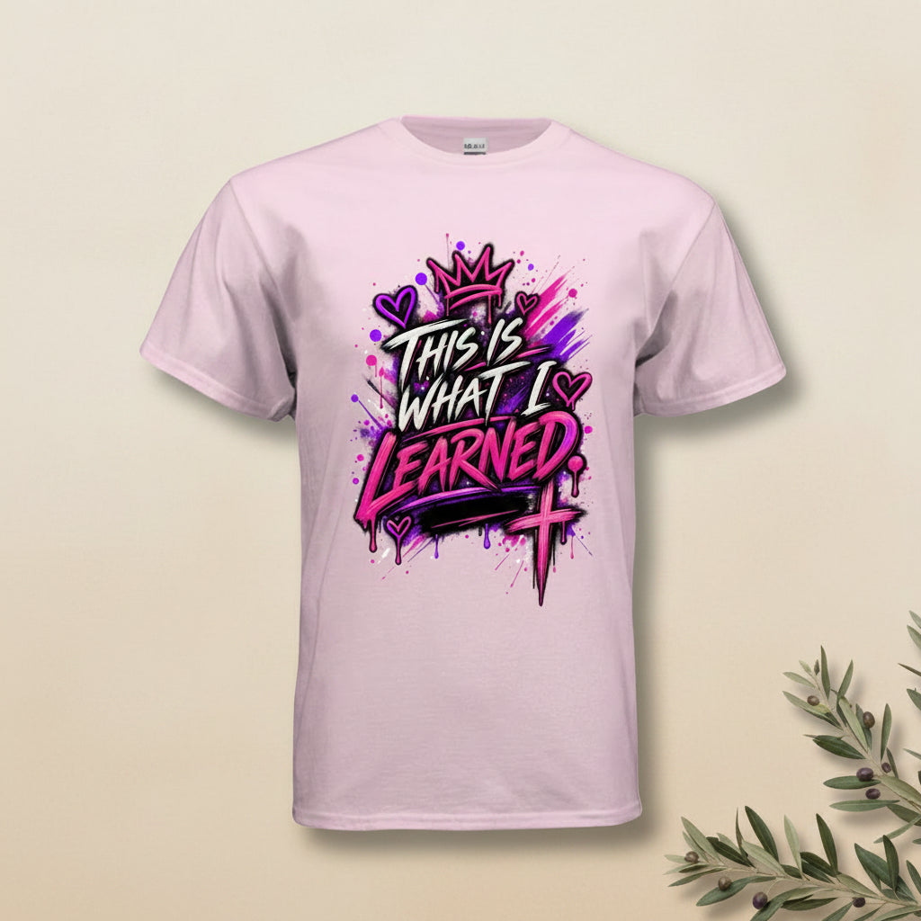 A light pink short-sleeve t-shirt with a colorful graphic on the front that reads 'THIS IS WHAT I LEARNED' in stylized black and pink lettering with purple and pink paint splatters and a crown.