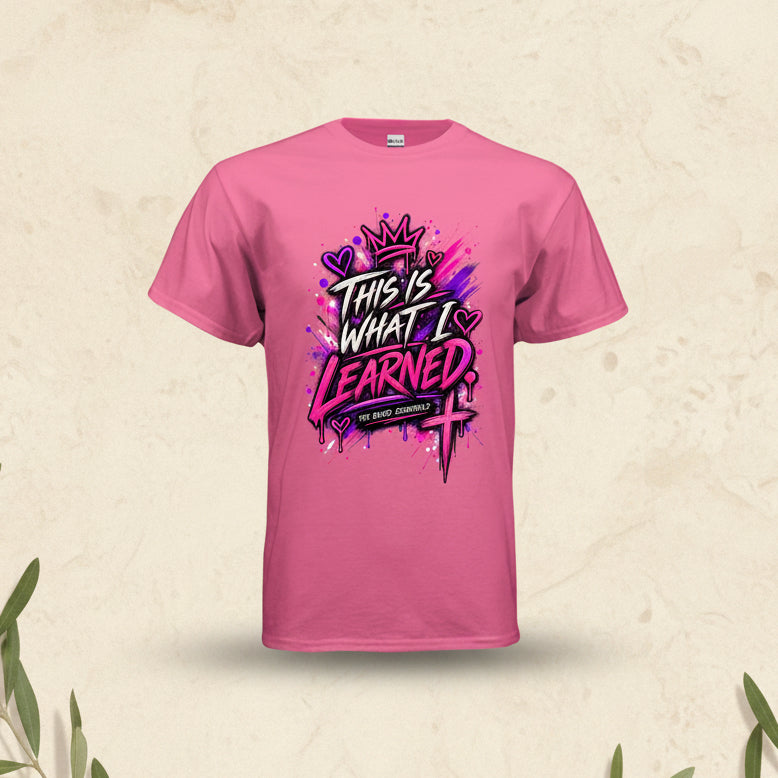 A pink short-sleeve t-shirt with a colorful graphic on the front that reads 'THIS IS WHAT I LEARNED' in stylized white, black, and pink lettering, with a crown and paint splatter design. The brand 'THE BATH ESSENTIALS' is visible on the graphic banner.