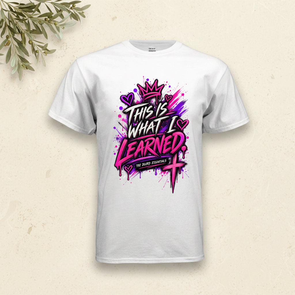 A white short-sleeve crewneck t-shirt featuring a colorful graphic print on the front. The graphic displays the text 'THIS IS WHAT I LEARNED' in a stylized black and pink font, decorated with a pink crown, purple hearts, and a pink cross, with 'THE BATH ESSENTIALS' written on a banner below.