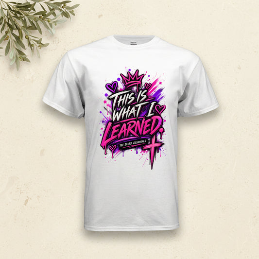 A white short-sleeve crewneck t-shirt featuring a colorful graphic print on the front. The graphic displays the text 'THIS IS WHAT I LEARNED' in a stylized black and pink font, decorated with a pink crown, purple hearts, and a pink cross, with 'THE BATH ESSENTIALS' written on a banner below.