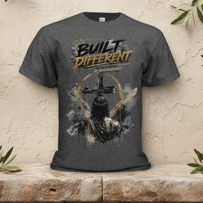 A dark heather gray graphic t-shirt featuring a distressed design with a cross, a halo, and the text 'BUILT DIFFERENT' in black and gold, displayed on a solid white background.