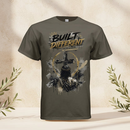 An olive green short-sleeve t-shirt with a distressed graphic on the front featuring a cross, a sword, and the text 'I AM BUILT DIFFERENT' in black and gold.